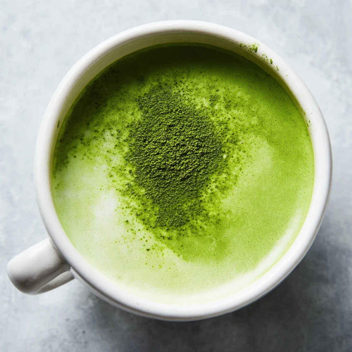 Vibrant green matcha latte with oat milk in a glass, topped with a thick layer of foam and a light dusting of matcha powder. 
