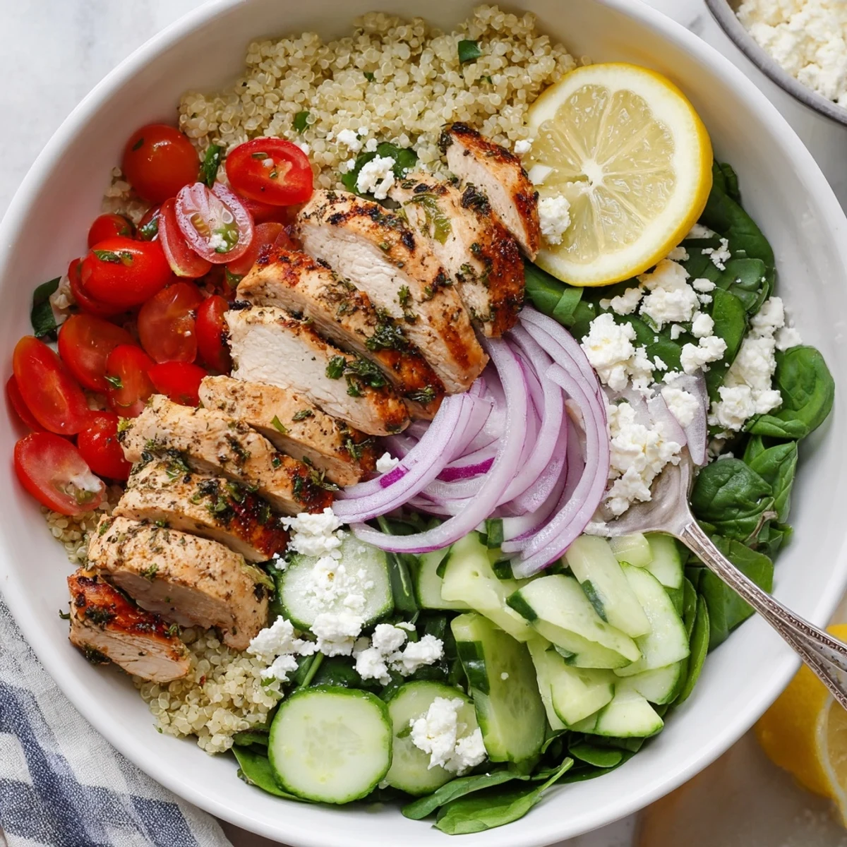 A close-up of Lemon Herb Grilled Chicken Quinoa Bowl with fresh herbs, feta, and lemon wedges ready to serve. 