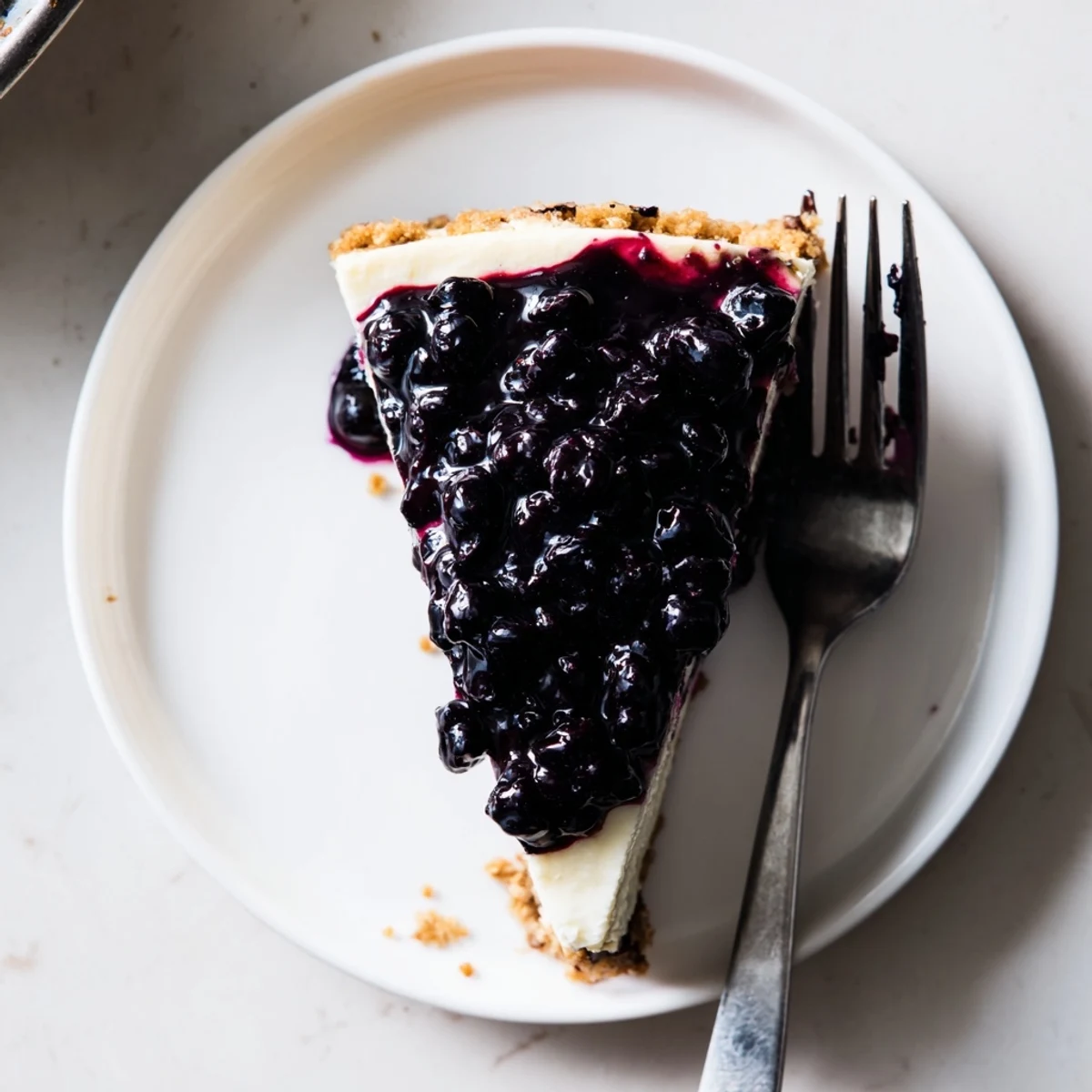 Creamy blueberry cheesecake slice topped with glossy purple compote on a white plate