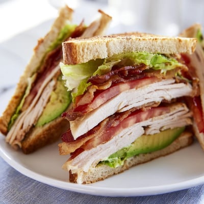 Triple-decker Turkey Club Sandwich with Turkey Bacon and Avocado, cut in half on a plate with a side of sweet potato fries.